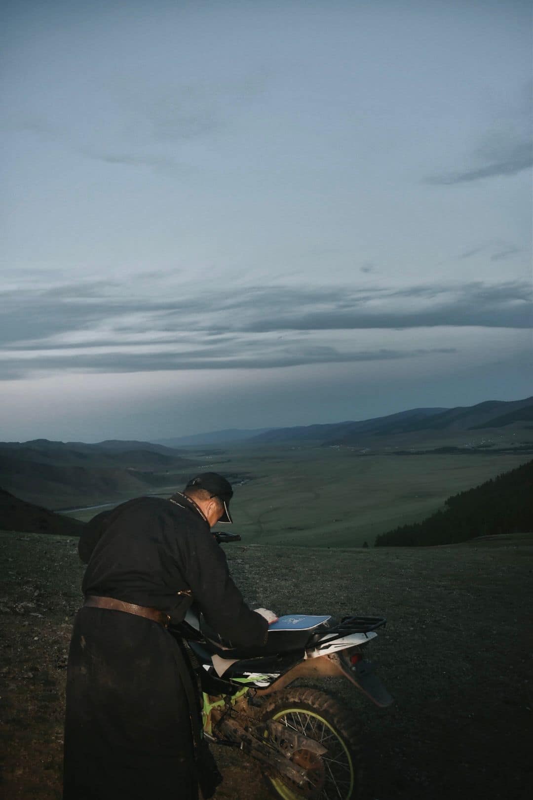 Herder in a deel loading his motorbike above the valley at dusk