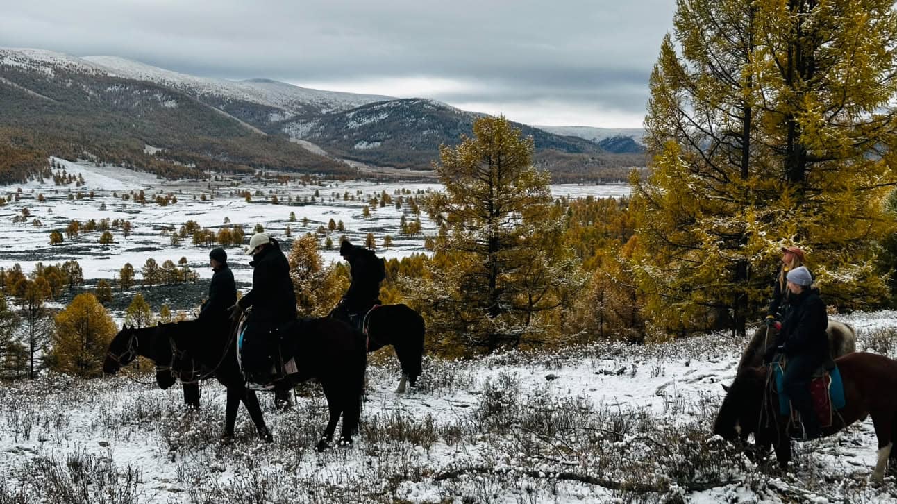 Riders pausing among golden larches after the first snow, the white valley and mountains beyond